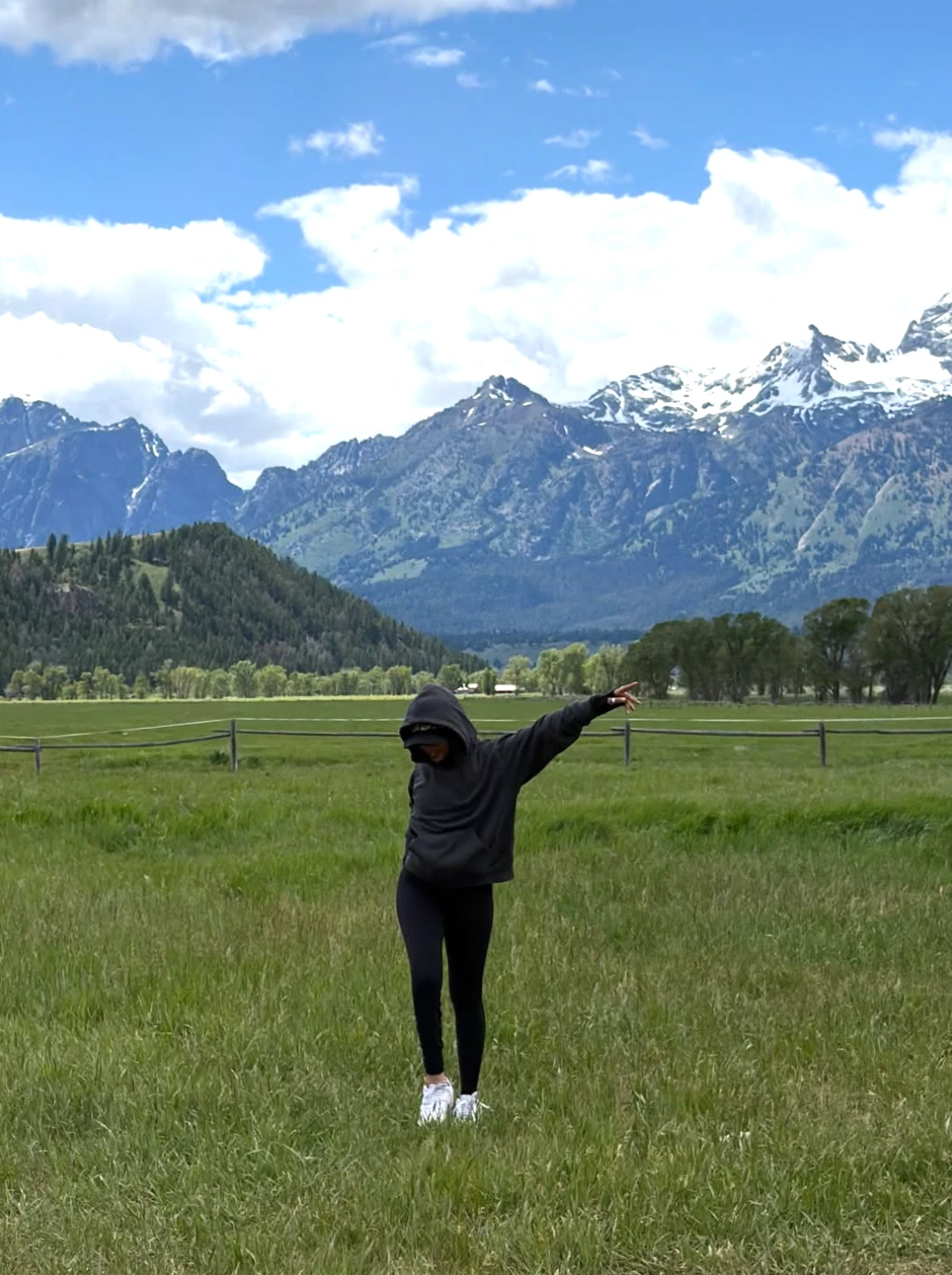 A person in a dark hoodie and leggings standing in a grassy field with one arm raised, facing a range of snow-capped mountains under a partly cloudy sky.